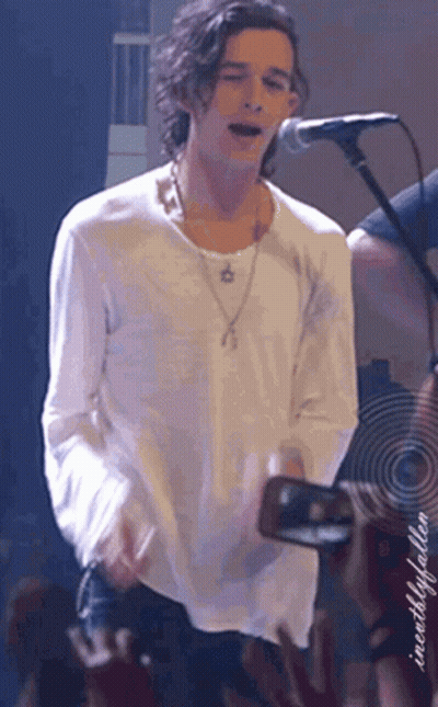 A Man In A White Shirt Is Singing Into A Microphone While Holding A Camera In Front Of A Crowd GIF