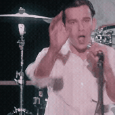 A Man In A White Shirt Is Singing Into A Microphone While Standing In Front Of A Drum Set . GIF