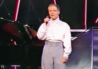 A Man In A White Shirt Is Singing Into A Microphone While Standing On A Stage GIF