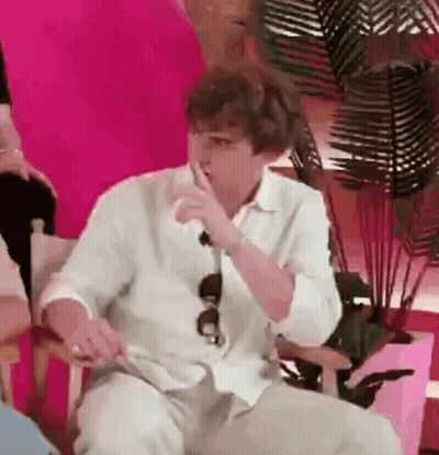 A Man In A White Shirt Is Sitting In A Chair With His Hand To His Mouth . GIF
