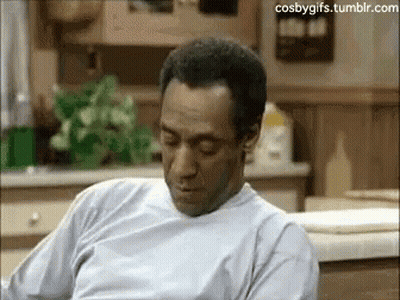 A Man In A White Shirt Is Sitting In A Kitchen Looking Down . GIF