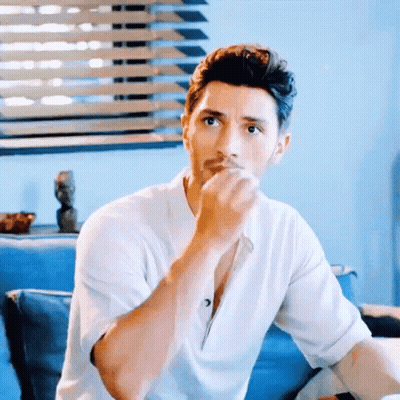 A Man In A White Shirt Is Sitting On A Blue Couch With His Hand On His Chin GIF
