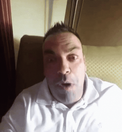 A Man In A White Shirt Is Sitting On A Couch And Making A Surprised Face . GIF