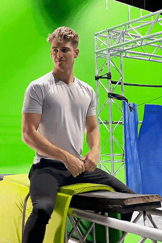 A Man In A White Shirt Is Sitting On A Yellow Cushion In Front Of A Green Screen GIF