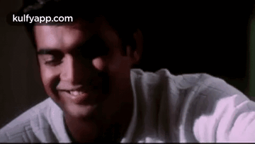 A Man In A White Shirt Is Smiling With His Eyes Closed In A Dark Room . GIF