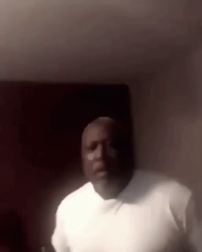 A Man In A White Shirt Is Standing In A Dark Room And Looking At The Camera . GIF