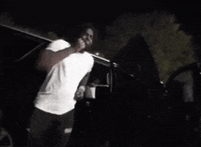 A Man In A White Shirt Is Standing In Front Of A Black Suv At Night . GIF