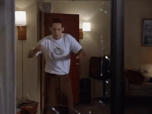 A Man In A White Shirt Is Standing In Front Of A Broken Glass Door . GIF