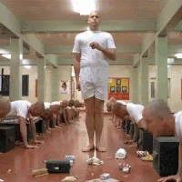 A Man In A White Shirt Is Standing In Front Of A Group Of Men Doing Push Ups On The Floor GIF