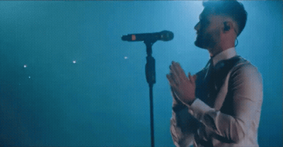 A Man In A White Shirt Is Standing In Front Of A Microphone Covering His Face . GIF