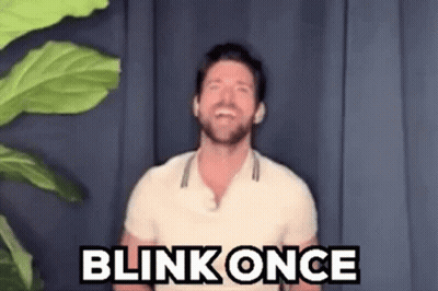 A Man In A White Shirt Is Standing In Front Of A Plant And Saying Blink Once . GIF