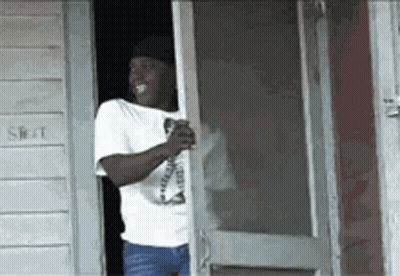 A Man In A White Shirt Is Standing In Front Of A Screen Door Holding A Drink . GIF