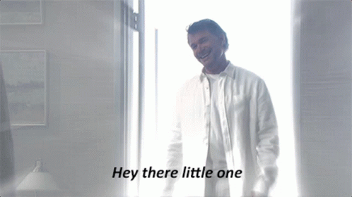 A Man In A White Shirt Is Standing In Front Of A Window With The Words Hey There Little One Below Him GIF