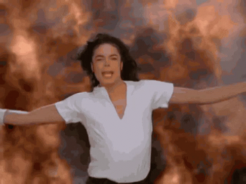A Man In A White Shirt Is Standing With His Arms Outstretched In Front Of A Fire Background GIF