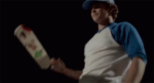 A Man In A White Shirt Is Swinging A Wooden Bat With A Green Heart On It GIF