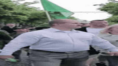 A Man In A White Shirt Is Walking Down A Street With A Green Flag In His Hand . GIF