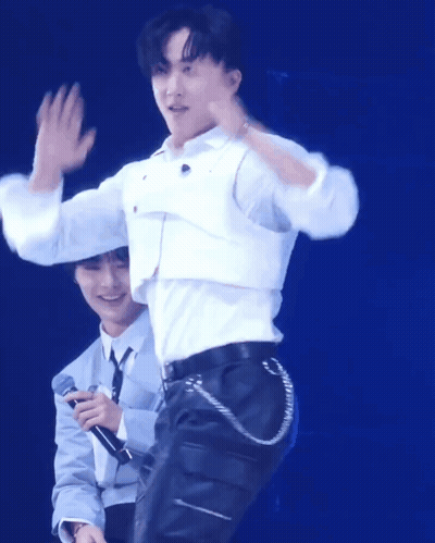 A Man In A White Shirt Is Waving While Another Man Holds A Microphone Behind Him GIF