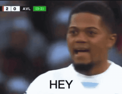 A Man In A White Shirt Says Hey In Front Of A Scoreboard GIF