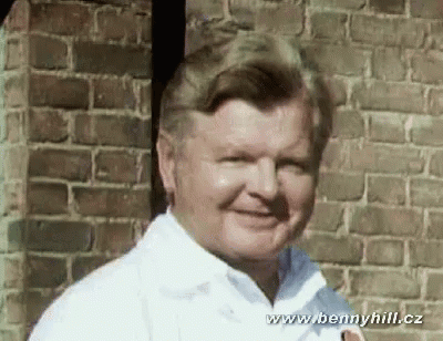 A Man In A White Shirt Stands In Front Of A Brick Wall With The Website Www.bennyhill.cz At The Bottom GIF