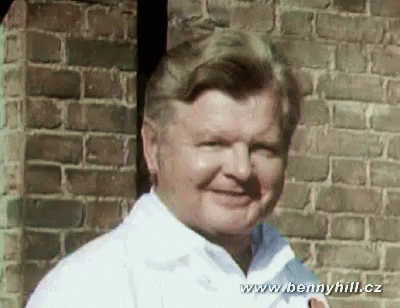 A Man In A White Shirt Stands In Front Of A Brick Wall With The Website Www.bennyhill.cz At The Bottom GIF