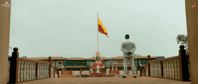A Man In A White Shirt Stands In Front Of A Building With A Flag Flying In The Background GIF