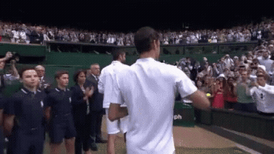 A Man In A White Shirt Stands In Front Of A Crowd Of People On A Tennis Court GIF