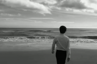 A Man In A White Shirt Stands On A Beach Looking At The Ocean GIF
