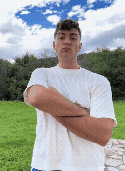 A Man In A White Shirt Stands With His Arms Crossed In A Field GIF