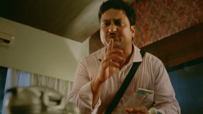 A Man In A White Shirt With A Black Strap Around His Shoulder Holds His Finger To His Lips GIF
