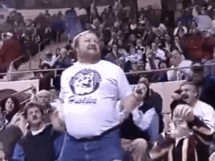 A Man In A White Shirt With A Lion On It Is Jumping In The Air In Front Of A Crowd . GIF