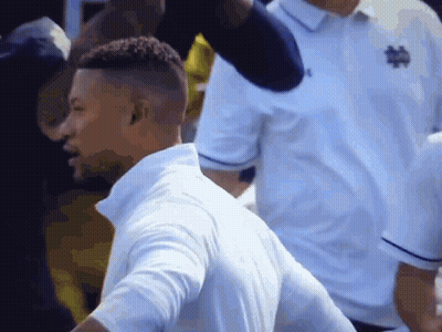 A Man In A White Shirt With A Notre Dame Logo On It Is Standing In A Crowd . GIF