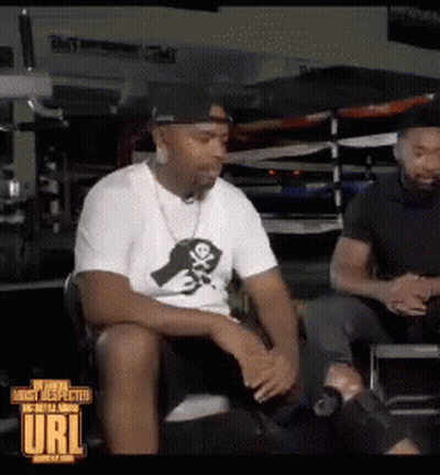 A Man In A White Shirt With A Skull And Crossbones On It Sits Next To Another Man In A Black Shirt GIF