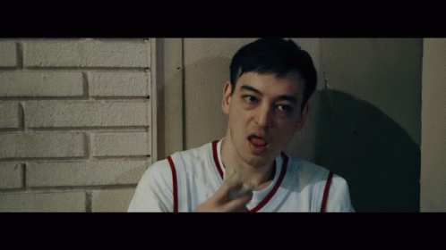A Man In A White Shirt With Red Stripes On The Sleeves Is Leaning Against A Brick Wall GIF