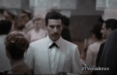 A Man In A White Suit And Black Bow Tie Is Walking Through A Crowd Of People . GIF
