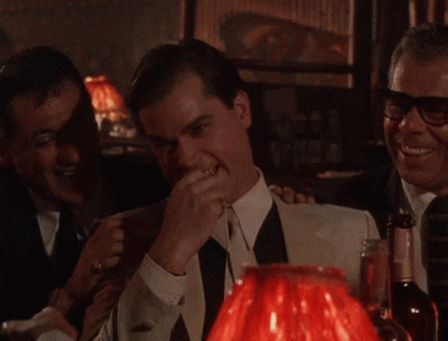 A Man In A White Suit And Black Tie Is Laughing In Front Of A Red Lamp GIF