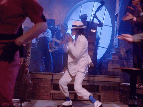 A Man In A White Suit And Blue Socks Is Dancing In Front Of A Group Of People GIF