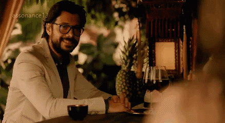 A Man In A White Suit And Glasses Is Sitting At A Table . GIF