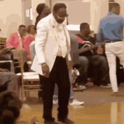 A Man In A White Suit Is Dancing In Front Of A Crowd Of People In A Room . GIF