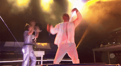 A Man In A White Suit Is Dancing On A Stage While A Woman Stands Behind Him With A Sign That Says Just Dance GIF