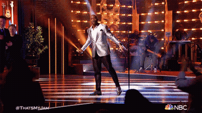a man in a white suit is singing into a microphone on a stage with a nbc logo in the corner