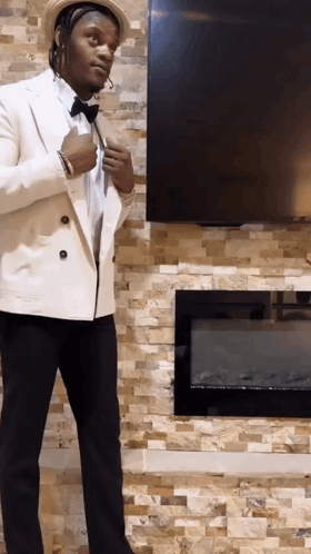 A Man In A White Suit Is Standing In Front Of A Fireplace And Television GIF