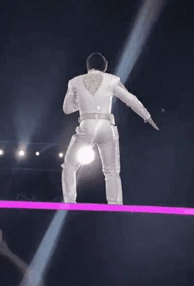 A Man In A White Suit Is Standing On A Stage With A Light Coming From His Back . GIF