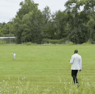 A Man In A White Suit Is Walking A Dog In A Field GIF