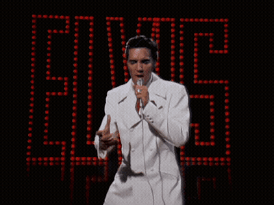 A Man In A White Suit Sings Into A Microphone In Front Of A Elvis Sign GIF