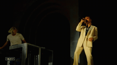 A Man In A White Suit Sings Into A Microphone While Another Man Plays A Keyboard In The Background GIF