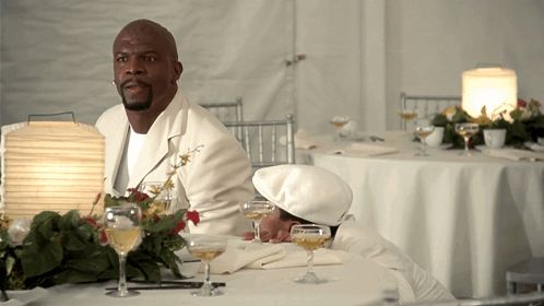 A Man In A White Suit Sits At A Table GIF