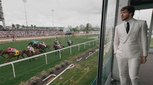 A Man In A White Suit Stands In Front Of A Horse Race GIF