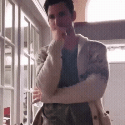 A Man In A White Sweater Is Standing In A Room With His Arms Crossed And His Hand On His Chin . GIF
