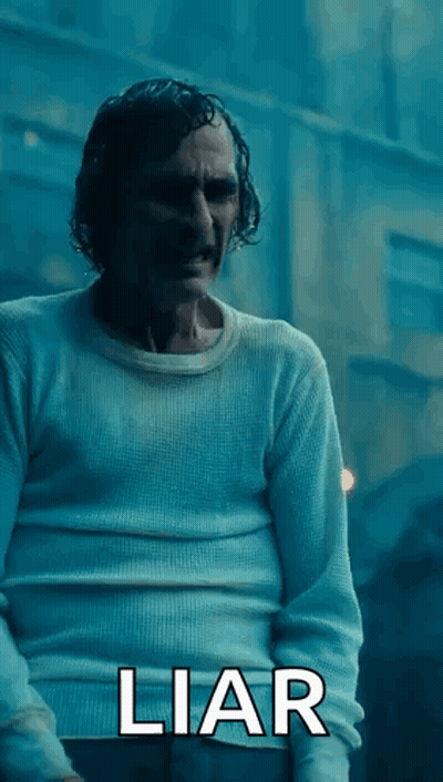 A Man In A White Sweater Is Standing In Front Of A Building With The Word Liar Written On The Bottom . GIF