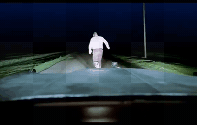 A Man In A White Sweater Is Walking Down A Road At Night GIF